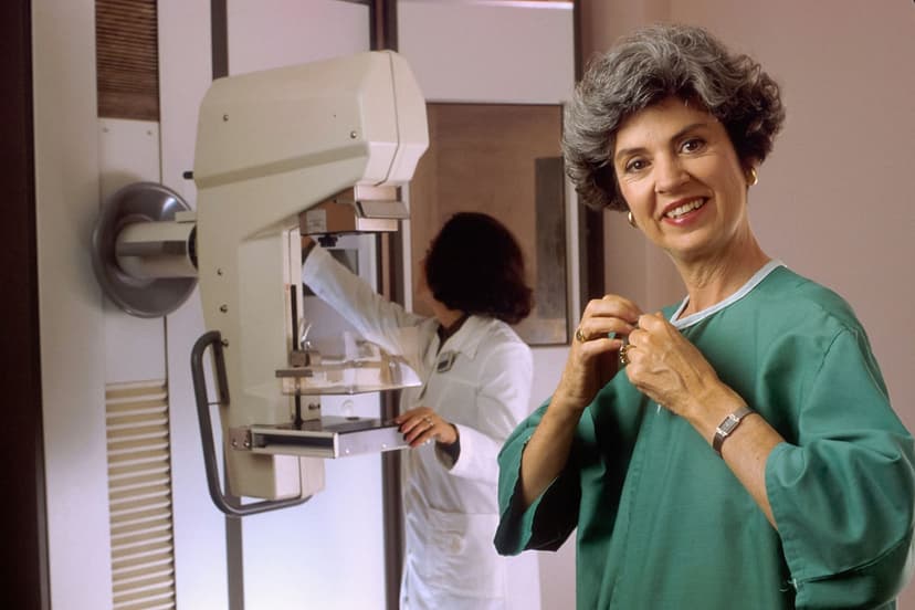 A woman in a hospital gown smiles beside a mammography machine as a technologist prepares it.